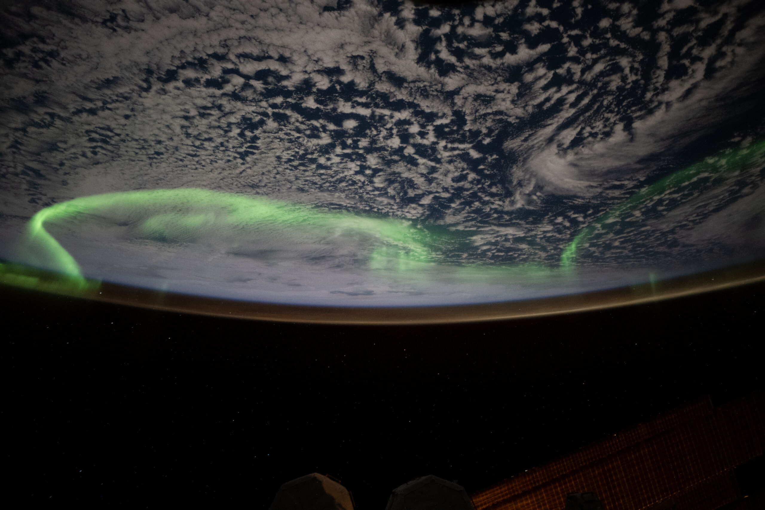 Green auroral arcs glow above clouds and Earth’s curved horizon in a photograph from the International Space Station.