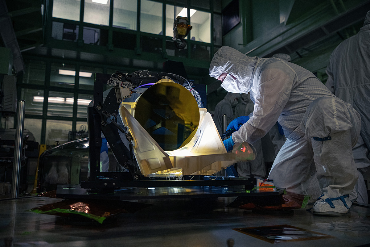 A clean-room technician inspects the gold-coated mirror of an amber-coloured LISA prototype telescope.