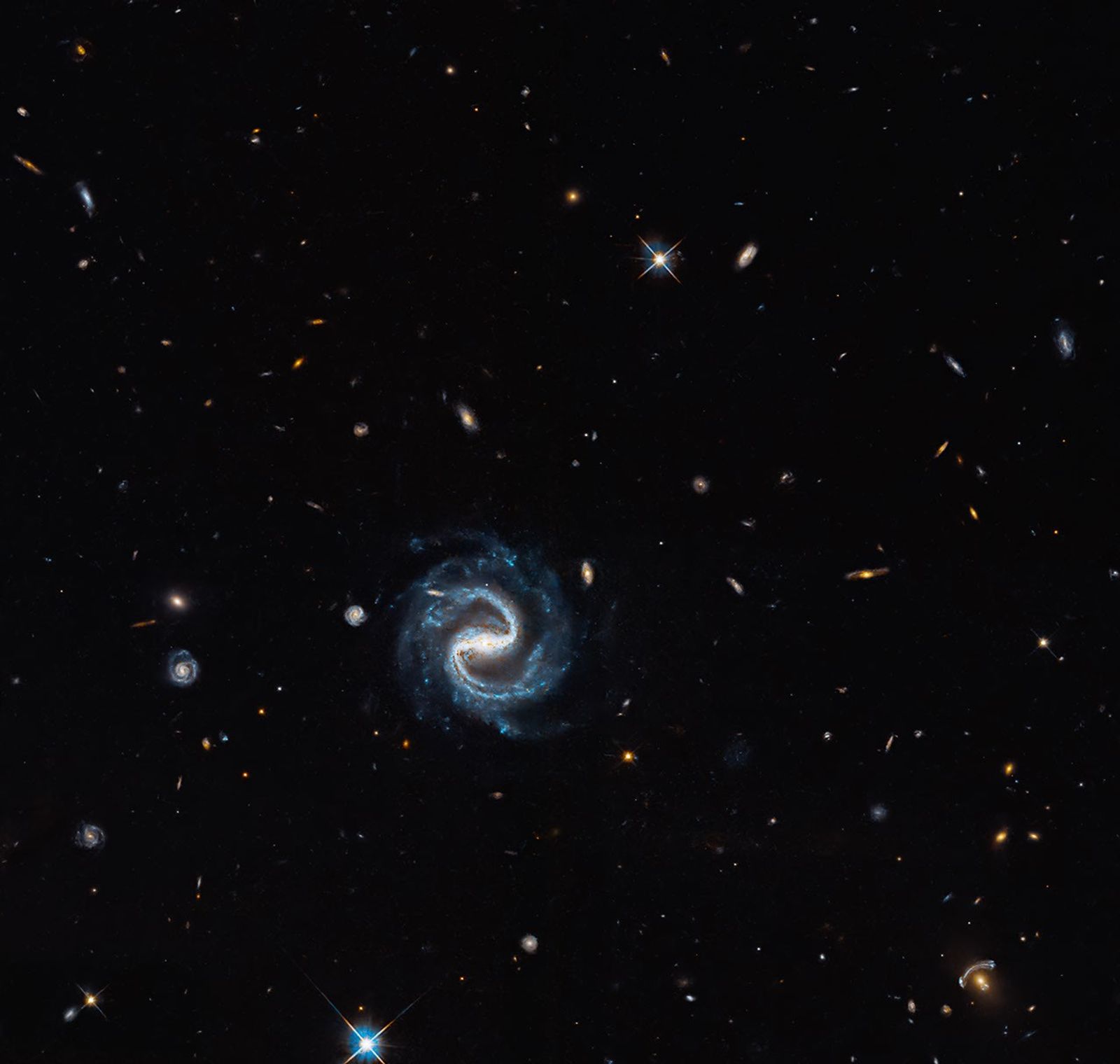 A blue barred spiral appears among distant galaxies; a golden galaxy and blue gravitationally lensed arc sit at lower right.