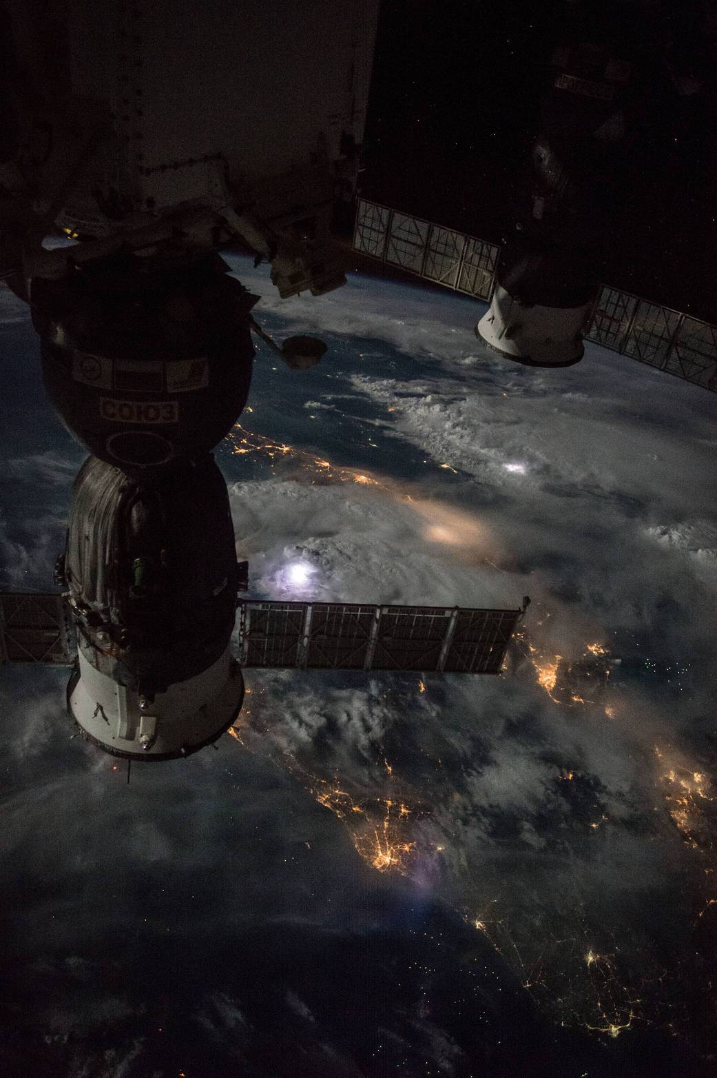 Bright lightning flashes illuminate clouds below docked Soyuz and Progress spacecraft in a nighttime view from the space station.