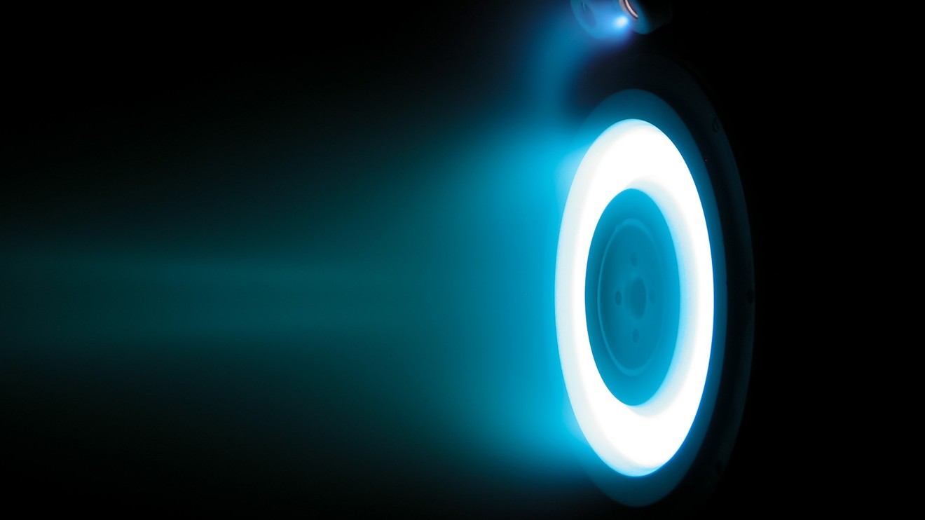 A bright blue ring and plume emerge from a Hall thruster inside a dark test chamber.