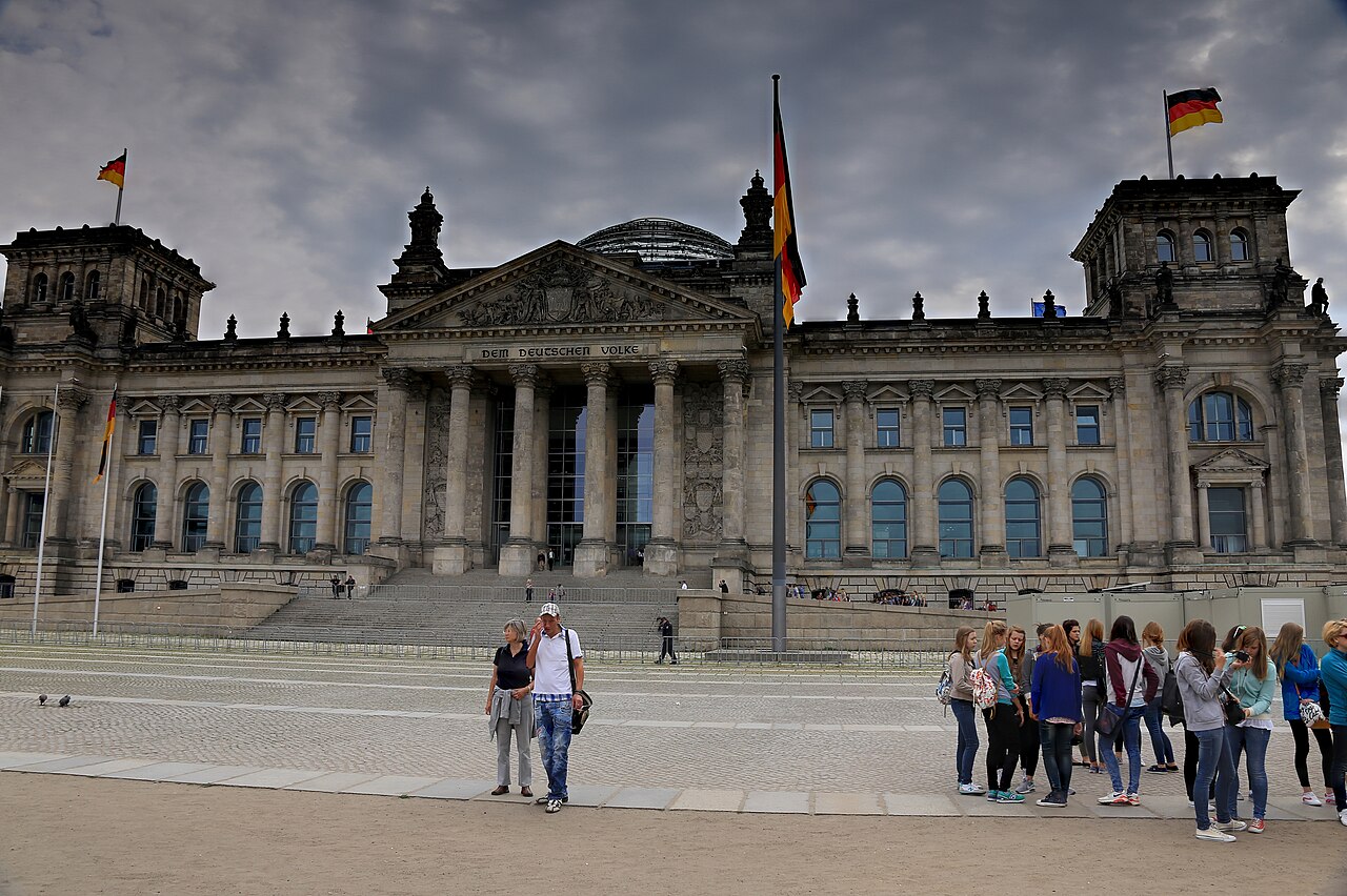 The Reichstag building in Berlin beneath gray clouds, with German flags and visitors in the foreground.