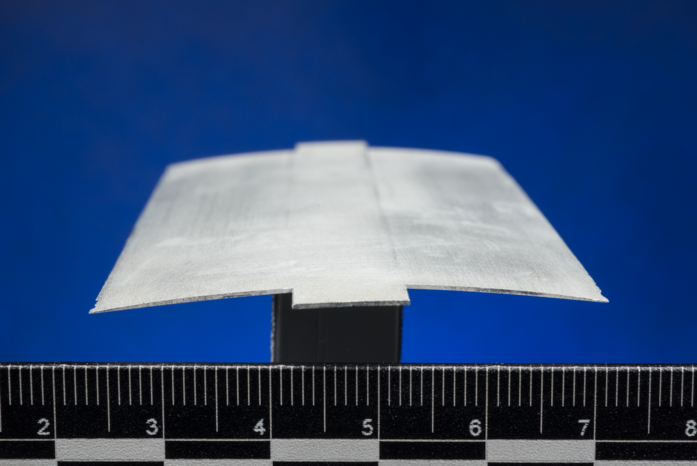A thin, gently curved gray airfoil model mounted on a black stand above a ruler against a blue background.