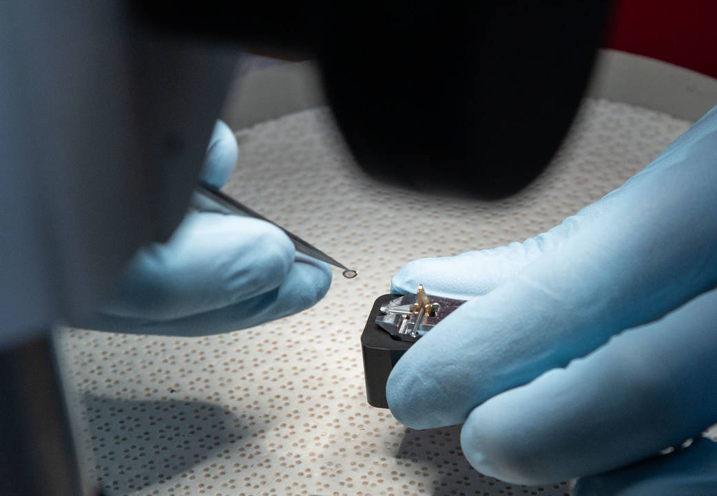 Gloved hands use a fine tool to handle a small asteroid Ryugu sample in a laboratory.